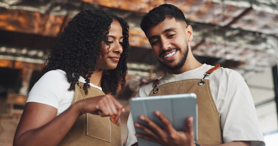 Two people in aprons smile while looking at a tablet, discussing work in a warmly lit space with a focus on teamwork.